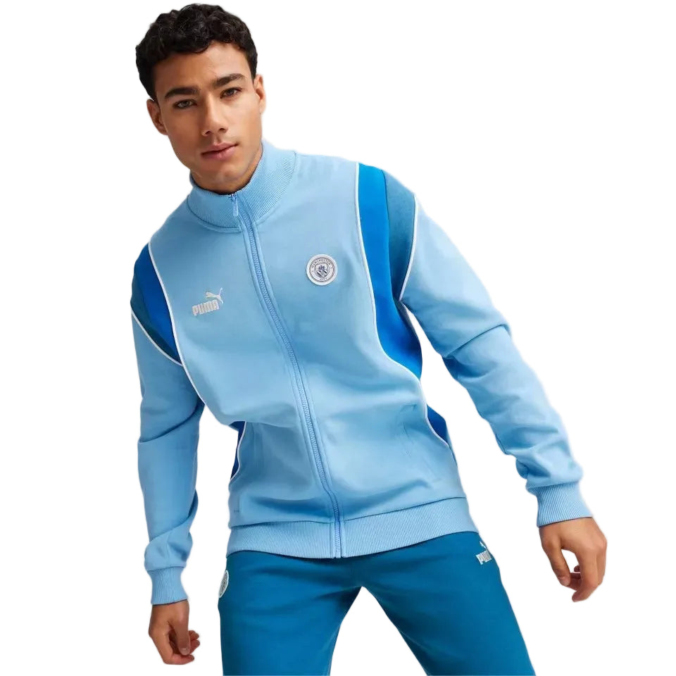 A man wearing a light blue Puma Manchester City track jacket with blue shoulder panels and matching blue track pants, posed against a light gray studio background.