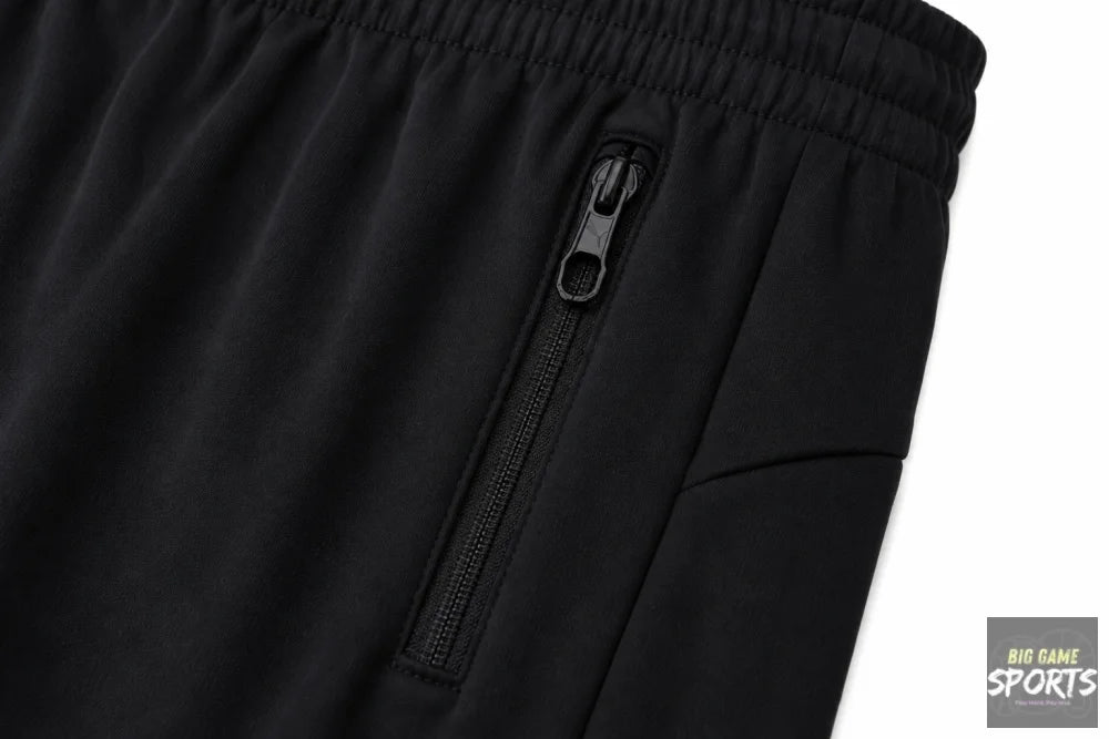 Close-up of black athletic pants showing a side zippered pocket with a black zipper pull and an elastic waistband, photographed against a white background.