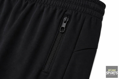 Close-up of black athletic pants showing a side zippered pocket with a black zipper pull and an elastic waistband, photographed against a white background.
