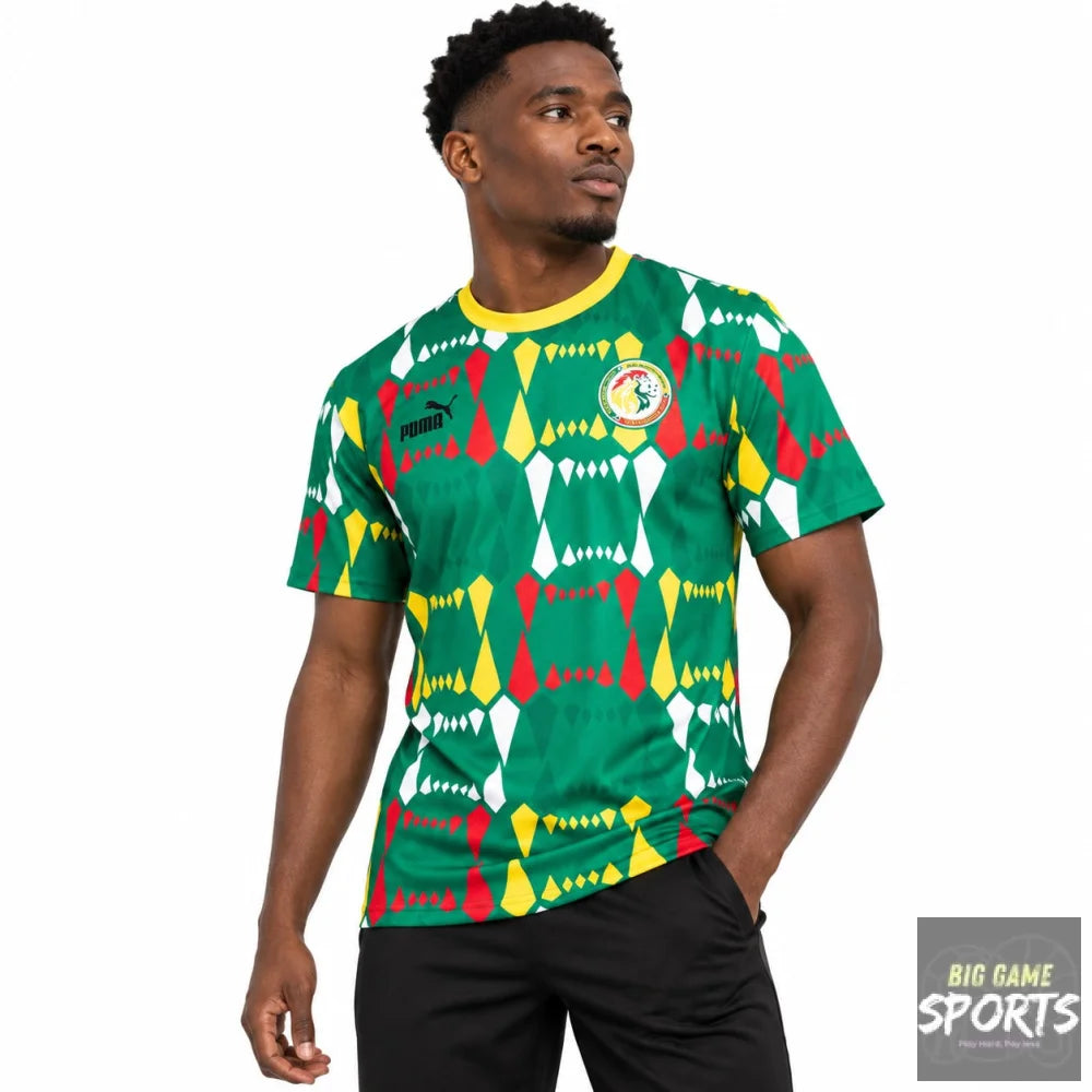 A man wearing a green short-sleeve soccer jersey with a colorful geometric pattern in red, yellow, and white, featuring a Puma logo and the Ghana national team crest on the chest.