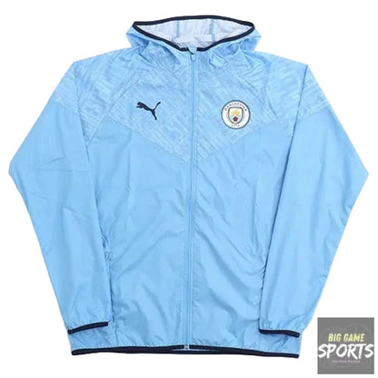 A light blue Puma Manchester City windbreaker jacket with a full zipper, featuring the Puma logo on the right chest and the Manchester City crest on the left chest, laid flat against a white background.