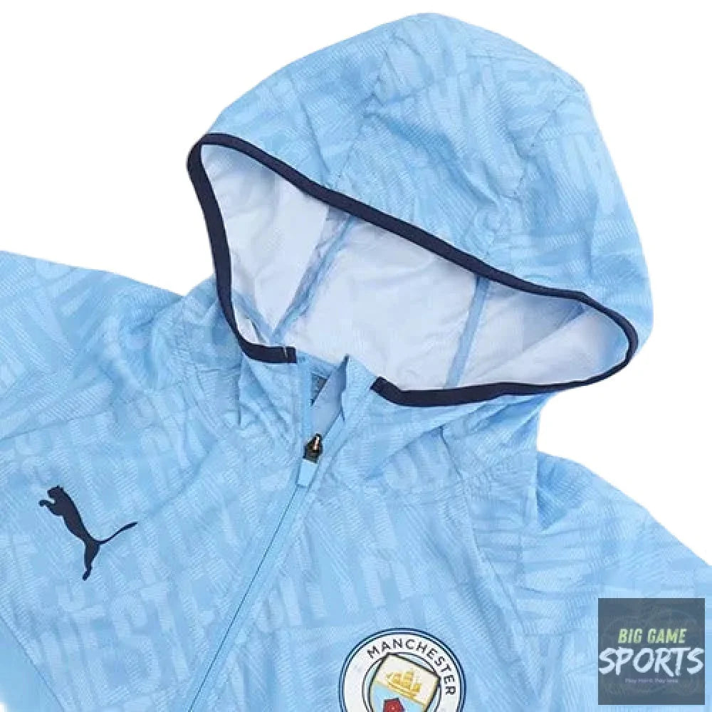 A light blue Manchester City Puma windbreaker jacket with a subtle geometric pattern, featuring a black Puma logo on the left chest and the Manchester City crest on the right chest. The hood is lined with a contrasting dark navy blue trim.