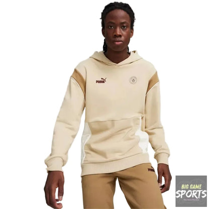 A model wearing a beige and tan Puma hoodie with a kangaroo pocket and matching tan sweatpants, standing against a light gray studio background.