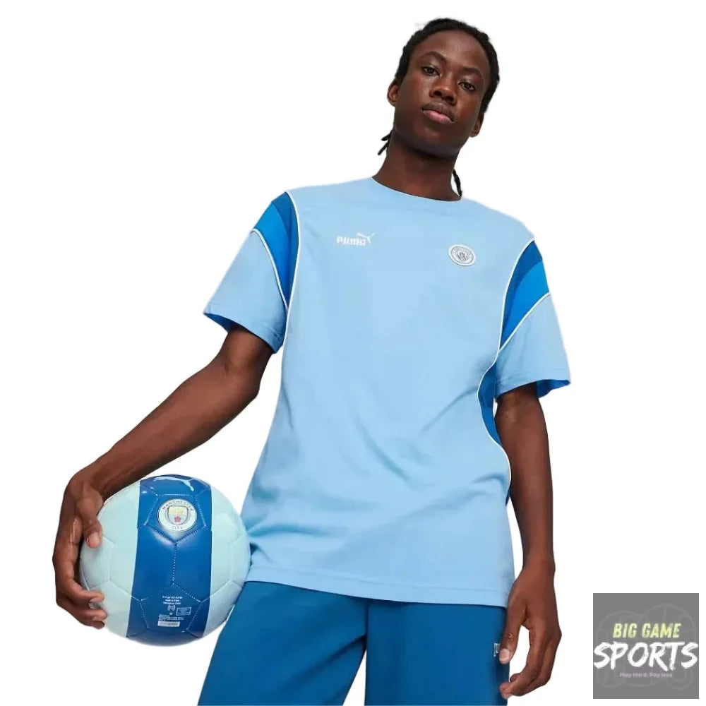 A model wearing a light blue and dark blue Puma soccer jersey with the Manchester City crest, holding a matching soccer ball against a light gray studio background.