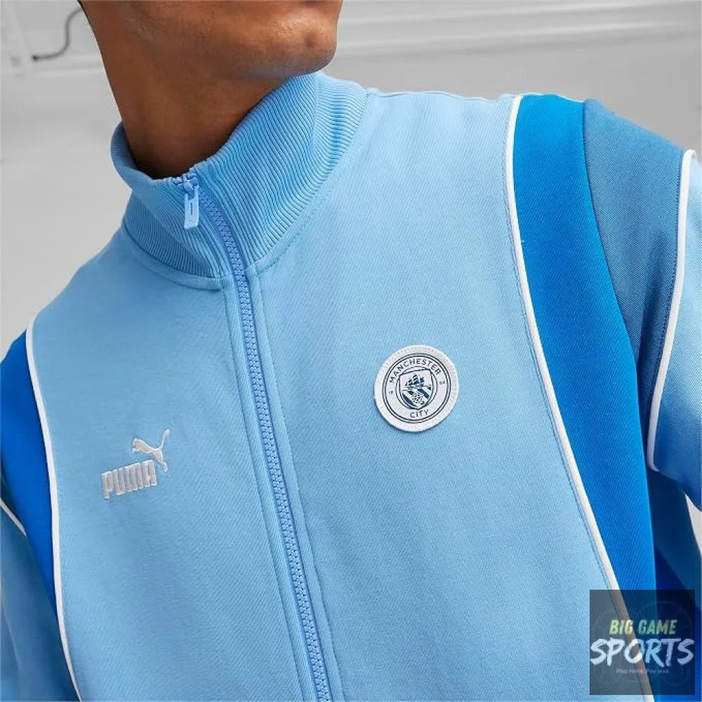A close-up of a person wearing a light blue Puma Manchester City track jacket, showing the front zipper, collar, Manchester City crest, and Puma logo.