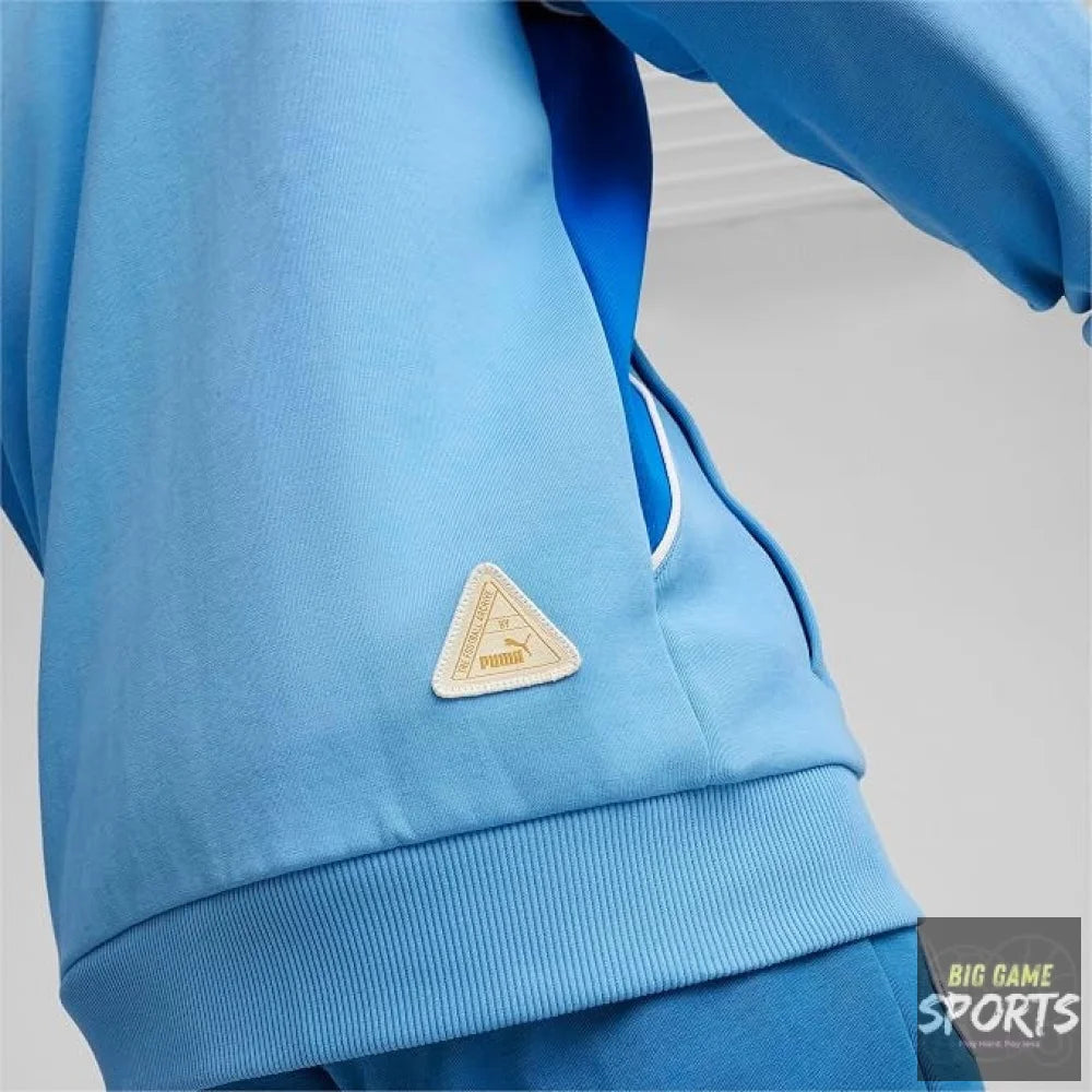 Close-up of a light blue Puma sweatshirt showing the ribbed hem, a side pocket with a white piping edge, and a beige triangular Puma logo patch.