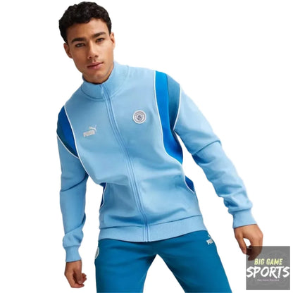 A man wearing a light blue Puma Manchester City track jacket with blue shoulder panels and matching blue track pants, posed against a light gray studio background.
