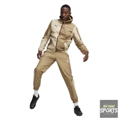 A model wearing a beige and cream Puma tracksuit with a hooded jacket and matching joggers, balancing a soccer ball on the floor in a studio setting.