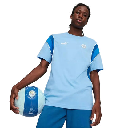 A model wearing a light blue and dark blue Puma soccer jersey with the Manchester City crest, holding a matching soccer ball against a light gray studio background.