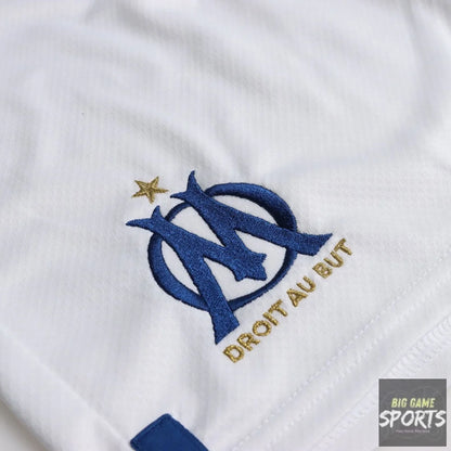 Close-up of a white soccer jersey fabric featuring the embroidered blue and gold Olympique de Marseille logo with the motto 'DROIT AU BUT' and a small gold star.