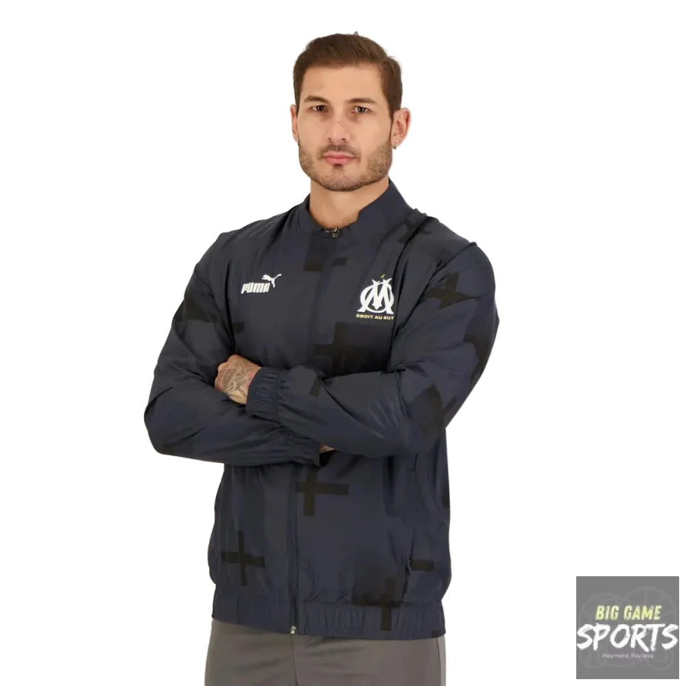 A man wearing a Puma Olympique de Marseille track jacket in navy blue with black star and cross patterns, posed against a light gray studio background.