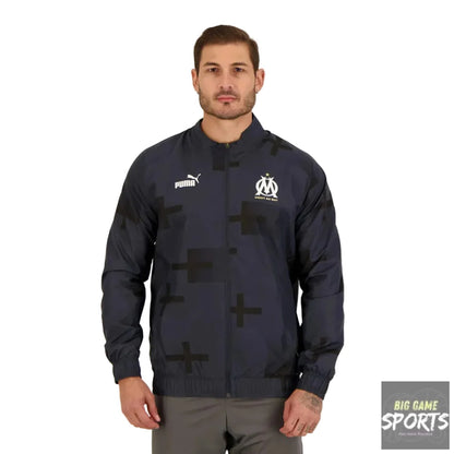 A man wearing a Puma Olympique de Marseille track jacket in navy blue with a black cross pattern, standing against a light gray studio background.