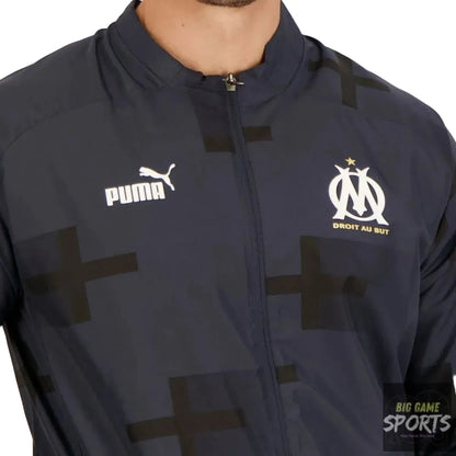 Close-up of a navy Puma track jacket with a subtle geometric cross pattern, featuring the white Puma logo on the right chest and the white Olympique de Marseille crest with gold text on the left chest.