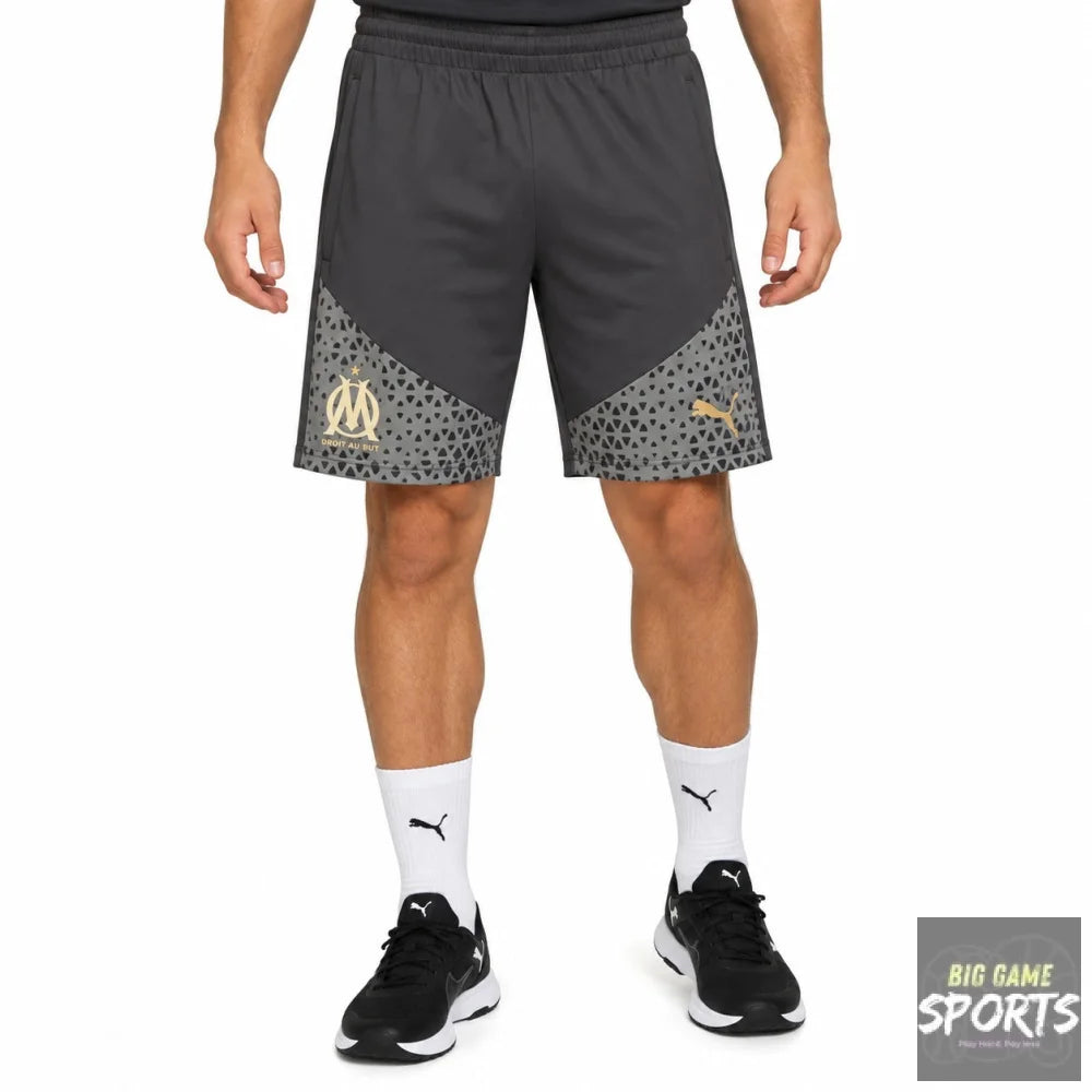 A model wearing dark gray Puma athletic shorts with a patterned lower panel and gold Olympique de Marseille crest, paired with white socks and black sneakers on a white studio background.