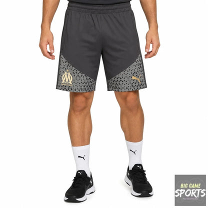 A model wearing dark gray Puma athletic shorts with a patterned lower panel and gold Olympique de Marseille crest, paired with white socks and black sneakers on a white studio background.