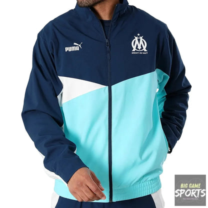 A man wearing a Puma Olympique de Marseille colorblock jacket in navy blue, light blue, and white, featuring the club's crest on the left chest and the Puma logo on the right.