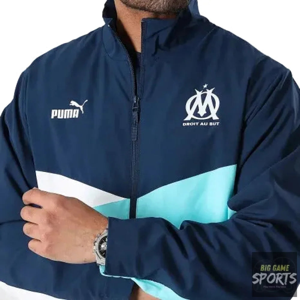 A close-up of a man wearing a Puma track jacket in navy blue with a white and light blue color-blocked design. The jacket features the Puma logo on the right chest and the Olympique de Marseille crest on the left chest.