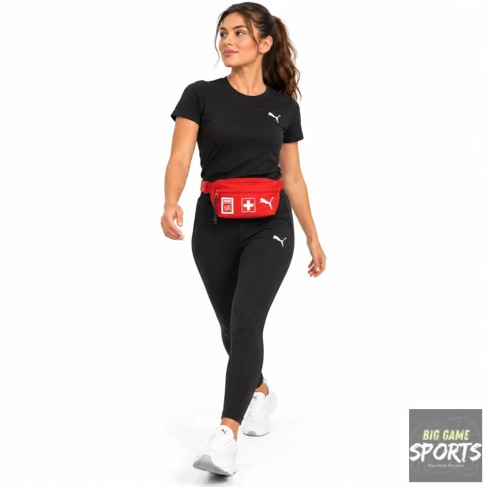 A woman modeling a red Puma fanny pack with a white Swiss cross logo, styled with a black Puma t-shirt and black leggings on a white studio background.