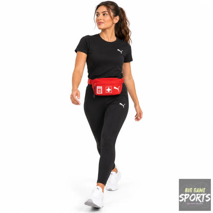 A woman modeling a red Puma fanny pack with a white Swiss cross logo, styled with a black Puma t-shirt and black leggings on a white studio background.