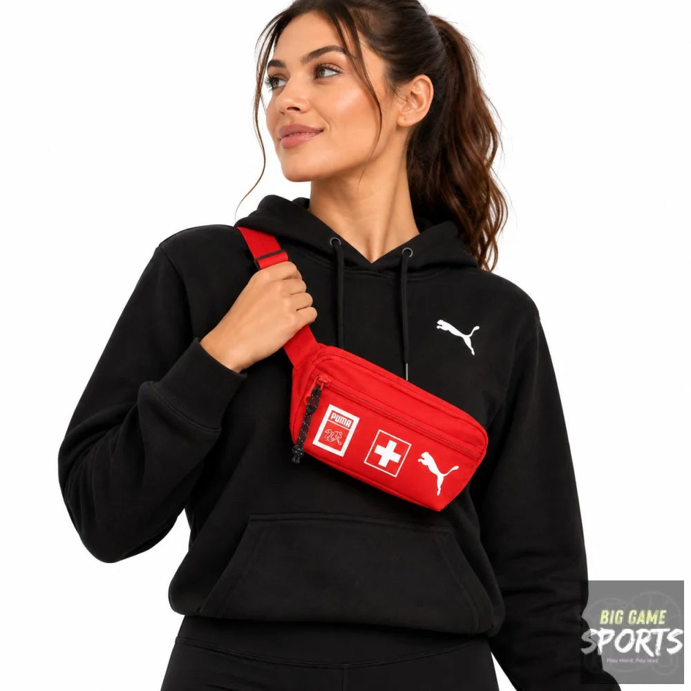 A woman wearing a black Puma hoodie and black sweatpants, carrying a red Puma Swiss flag fanny pack on her shoulder against a white background.