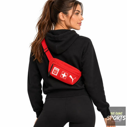 A woman wearing a black hoodie and black leggings from the back, wearing a red Puma crossbody bag with Swiss flag graphics on the front, against a solid white background.